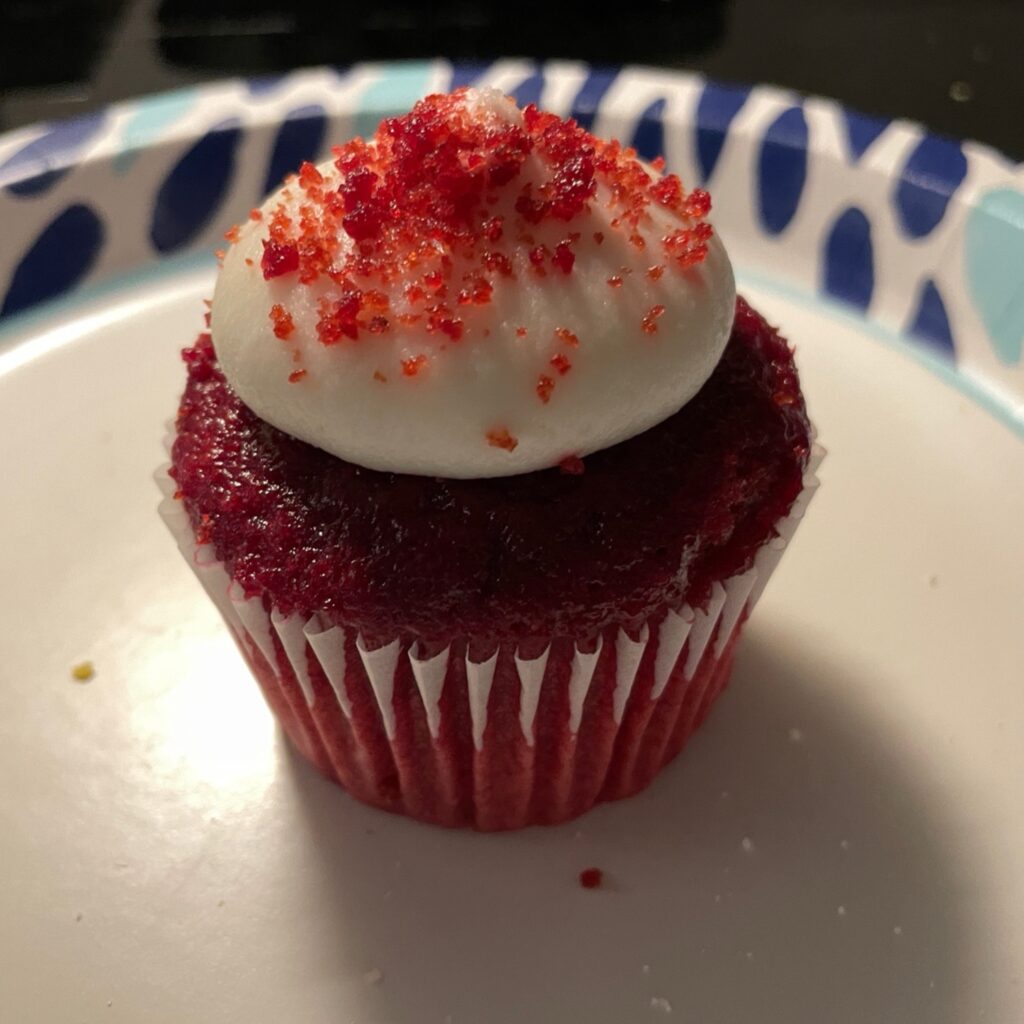 A red velvet cupcake with a dollop of cream cheese frosting and red crumbles on top. 