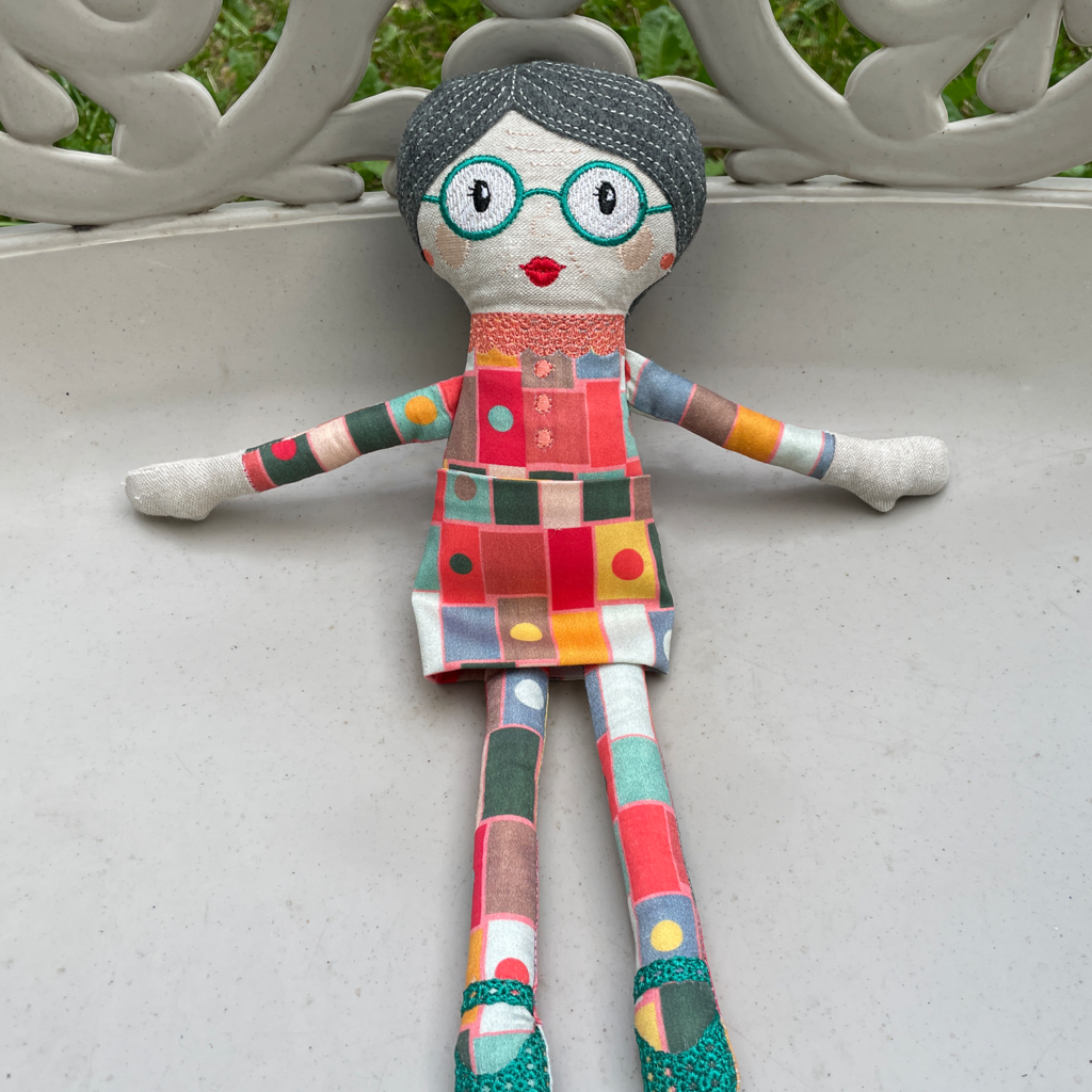 A grandma doll with grey hair, turquoise eyeglasses, and a mini skirt sitting on a lawn chair. 