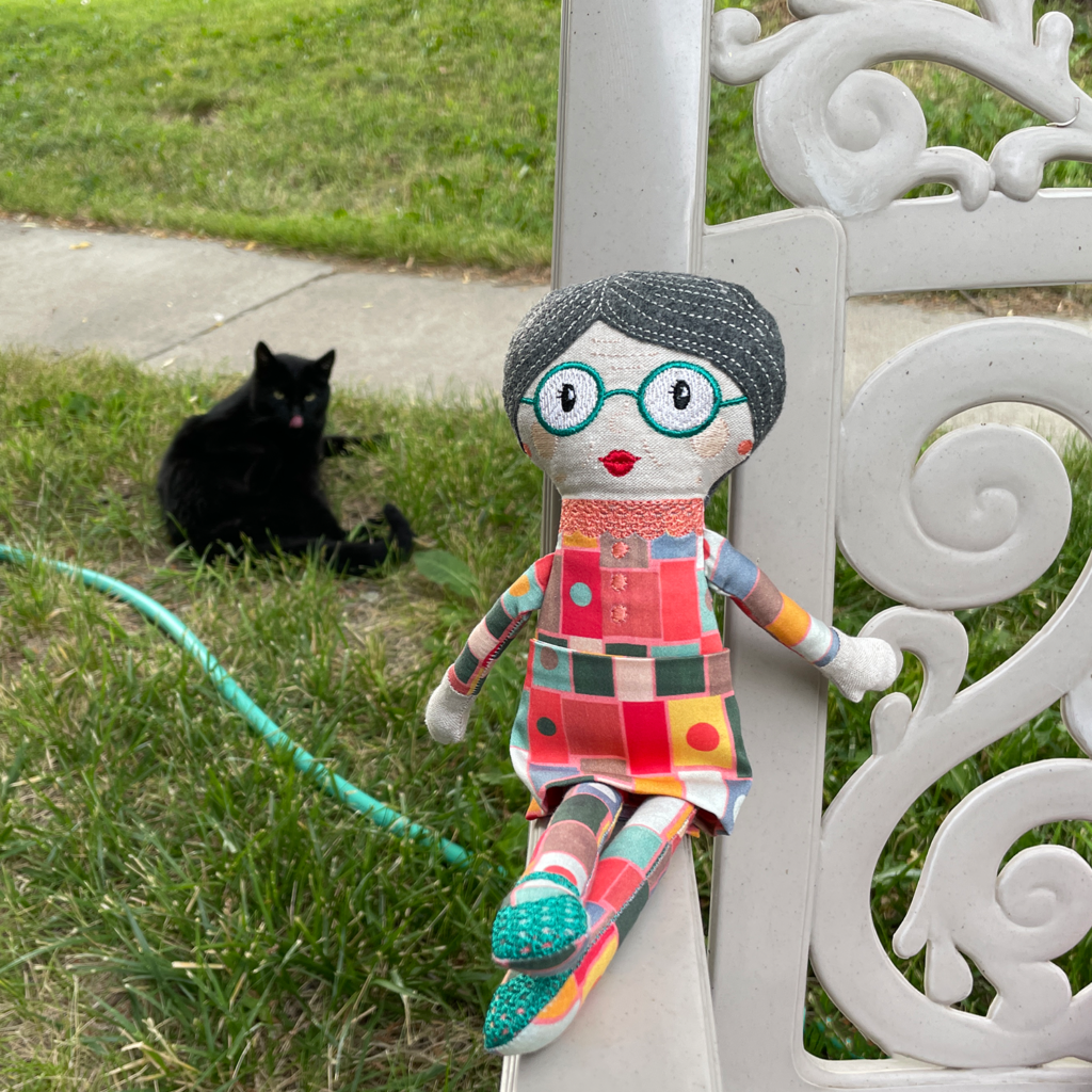 The Grandma doll I sewed is sitting on the arm of a plastic lawn chair. Jet the kitty is in the background with his tongue sticking out. 