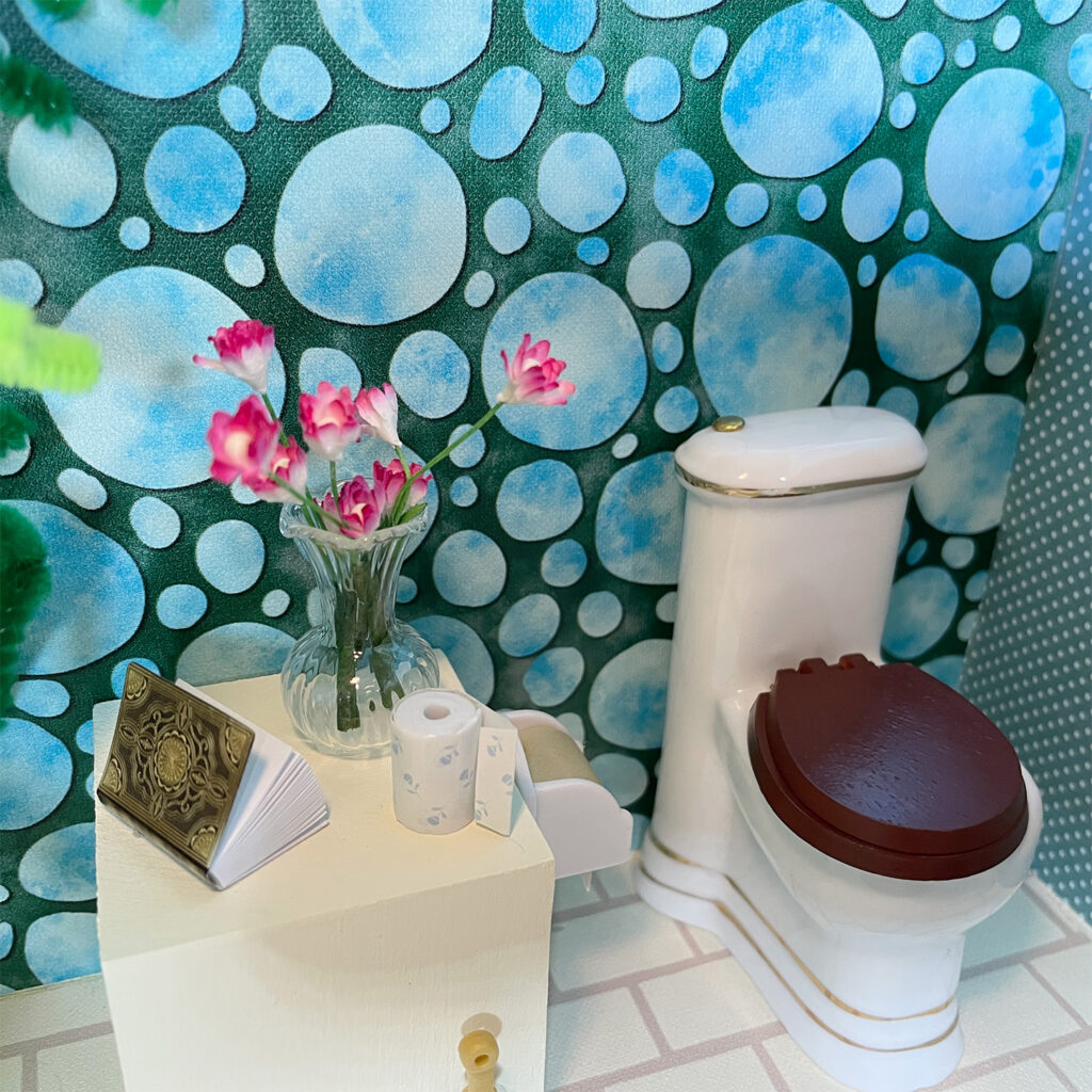 A table beside the toilet has a bouquet of flowers, a spare roll of toilet paper, and a book. 