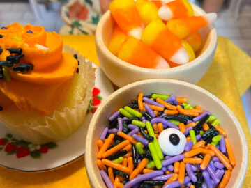 A cupcake on a plate, a mini bowl of candy corn, and a mini bowl of candy worms and an eyeball.