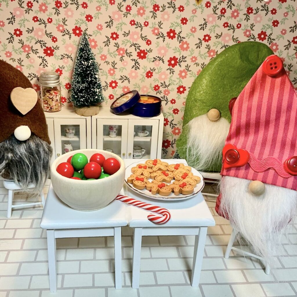 Three gnomes around a table with a bowl of gumboils, a plate of gingerbread cookies, and a candy cane. 