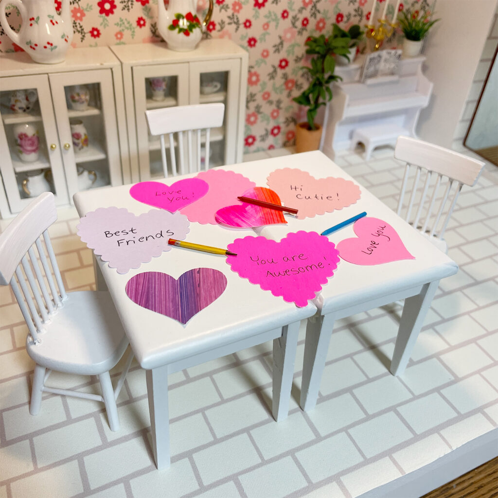 The dollhouse dining room table is covered with heart-shaped valentines, pencils, and crayons. 