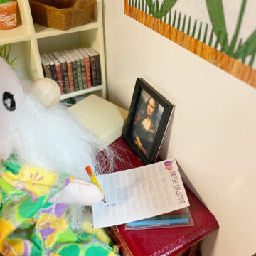 A little doll with a beard and a bead nose is sitting at a desk writing on a piece of paper.